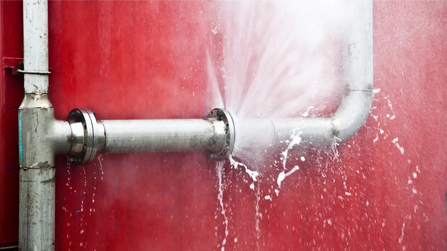 Burst metal pipe spraying water against red wall – water damage restoration in Tustin